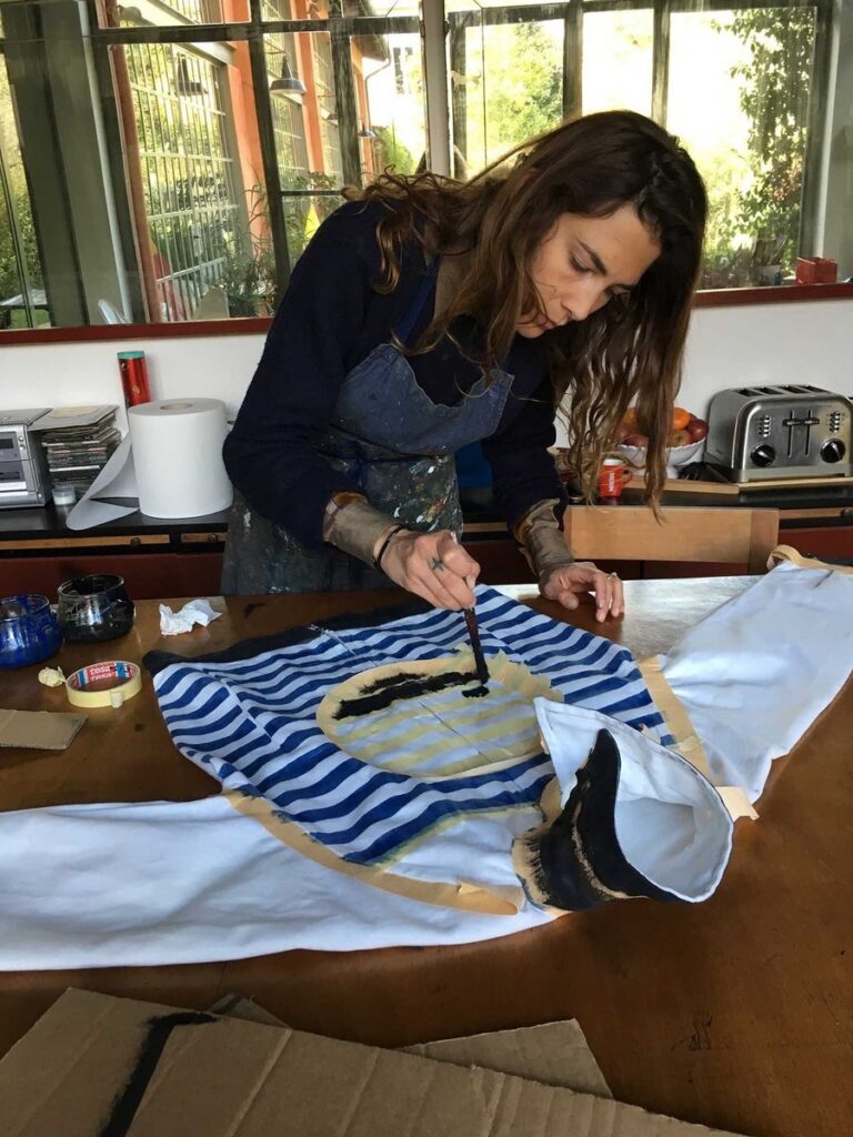 Zazie peignant au pinceau un motif sur un tissu rayé bleu et blanc tendu sur une table de l'atelier, lumière naturelle des grandes fenêtres ouvertes sur le jardin.