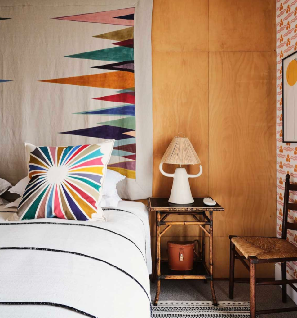 Chambre avec coussin sunburst et tenture murale motif Raggiera (éventails de couleurs vives sur fond clair), lit blanc à rayures, lampe sur table de chevet en bois.