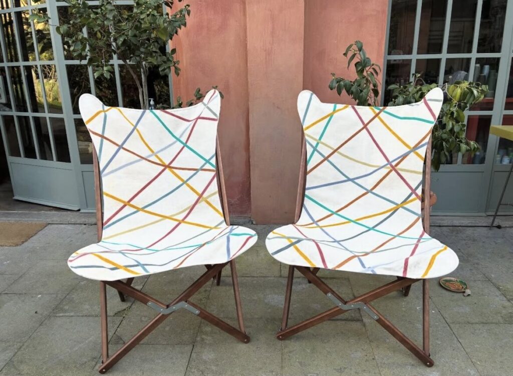 Deux chaises pliantes en bois, sièges et dossiers retendus avec un tissu blanc peint à la main de lignes croisées multicolores.