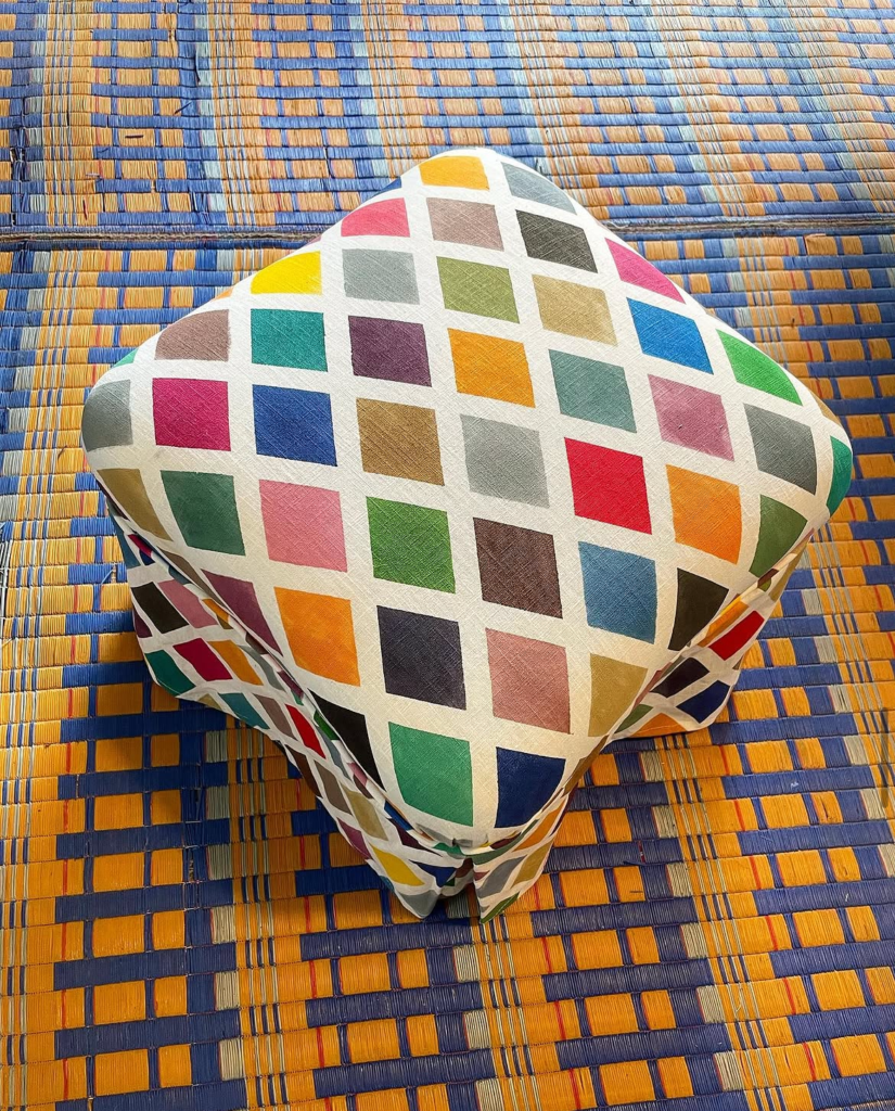 Coussin pouf carré rebondi recouvert du motif Checks (mosaïque de carrés multicolores), posé sur une natte tressée bleue et orange.