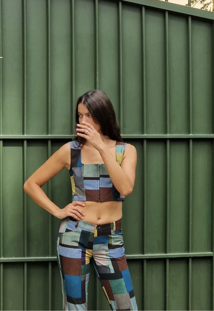 Mannequin portant un ensemble bustier et pantalon coupé dans un tissu patchwork de damiers bleu, brun et turquoise, photographié devant une grille métallique vert sapin.
