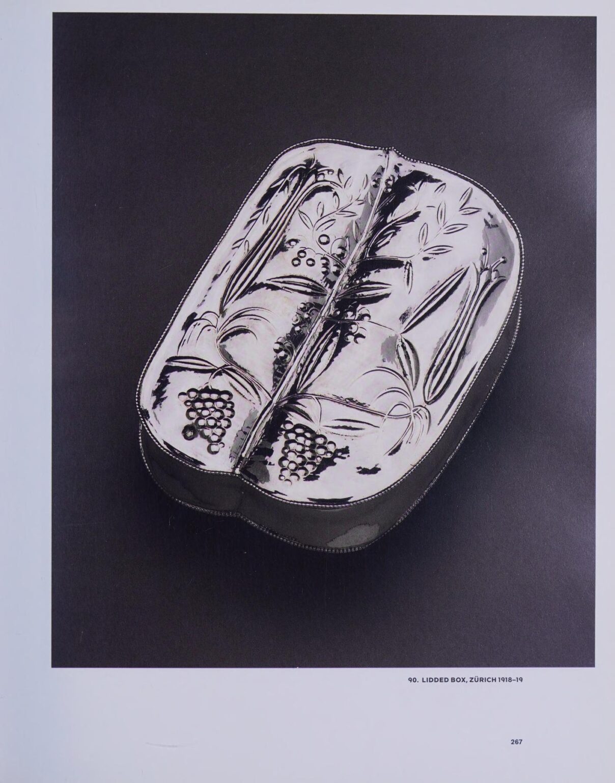 Silver lidded box with chased decoration of grape vines and foliage, photographed at an angle on a black background.