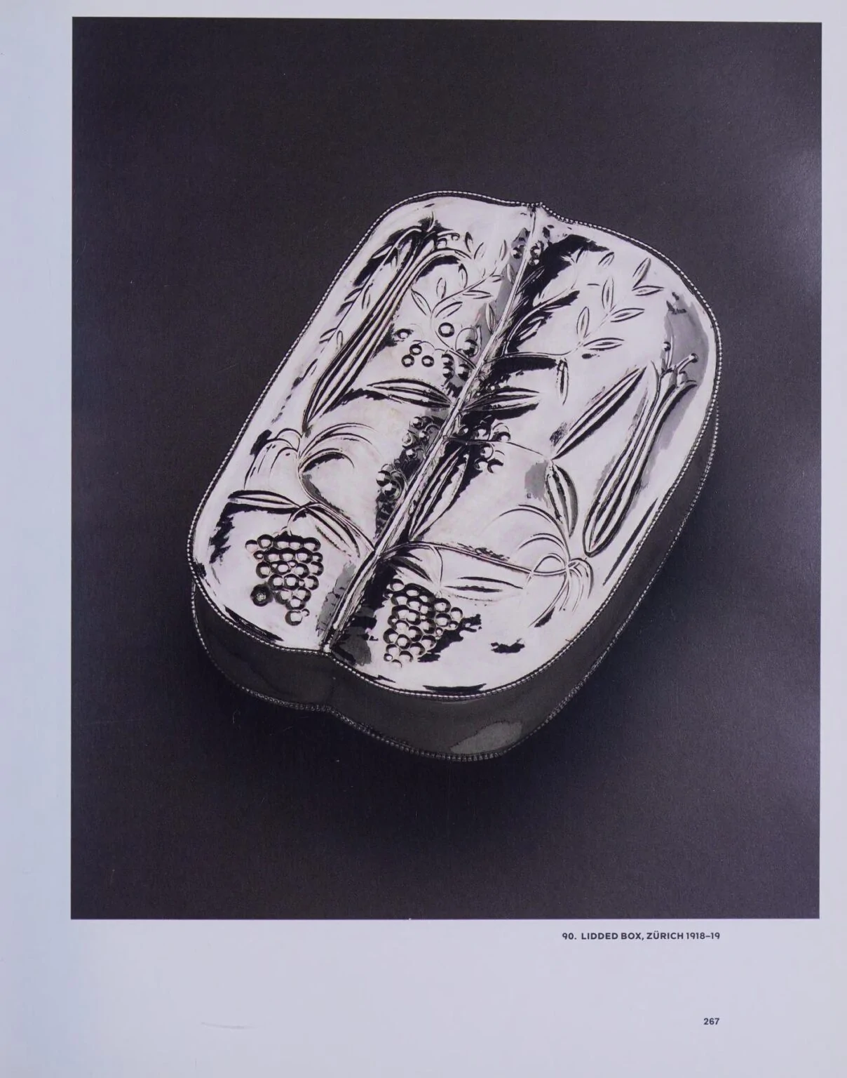 Silver lidded box with chased decoration of grape vines and foliage, photographed at an angle on a black background.