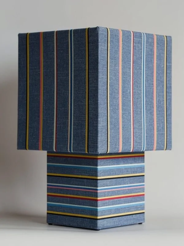 Lampe de table cubique entièrement habillée du même métrage rayé bleu denim et fils colorés, abat-jour et fût recouverts du même tissu.
